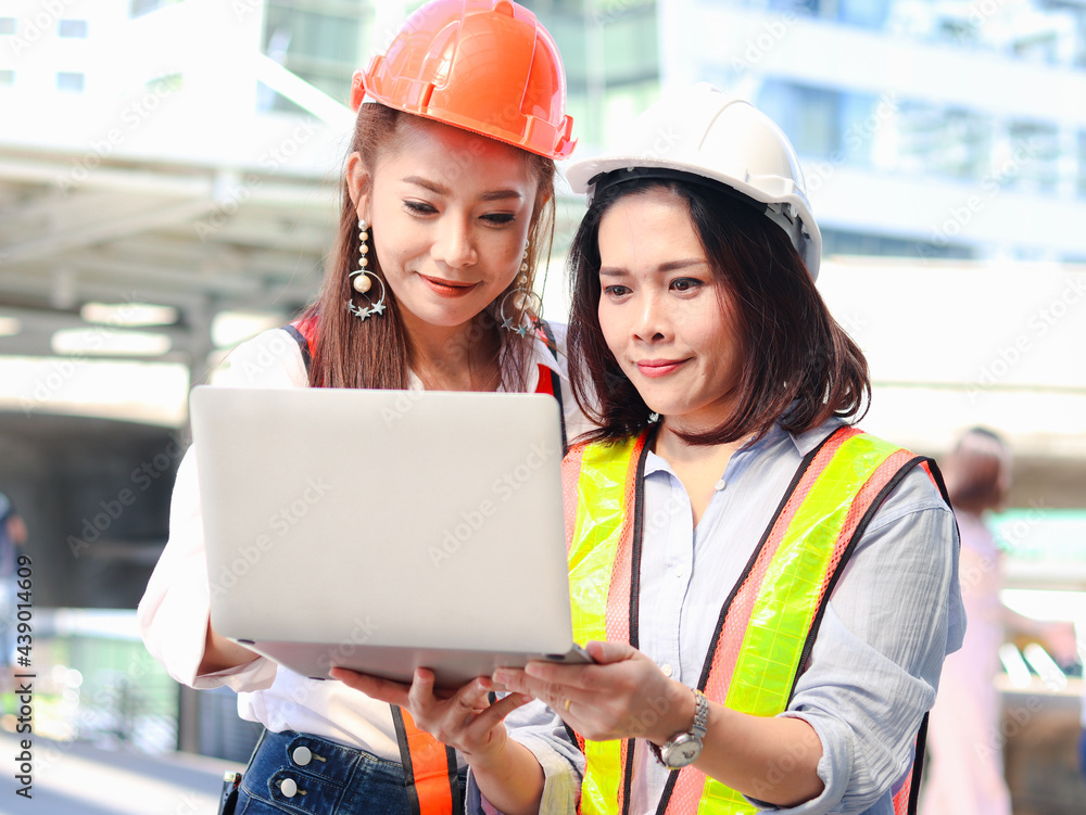 Two young beautiful Asian female engineer workers wearing safety vest and helmet, using laptop computer together, pretty women discussing about project work while walking on the way outside office in Two young beautiful Asian female engineer workers wearing safety vest and helmet, using laptop computer together, pretty women discussing about project work while walking on the way outside office in