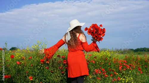 beautiful curly-haired woman in a red dress in a white straw hat walks along a poppy field with a bouquet of wildflowers, standing with her back