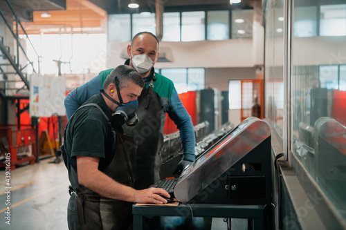 two workers wearing a face mask due to a coronavirus pandemic are programming a modern cnc machine