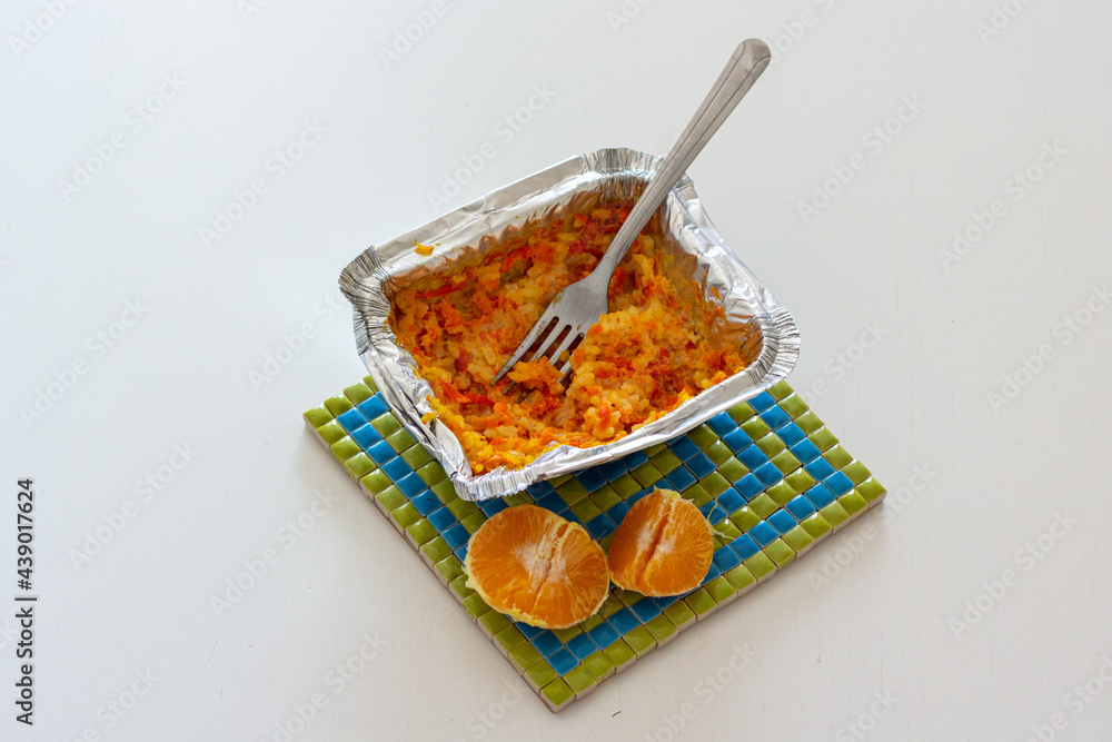 Fototapeta premium Carrot rice porridge in foil packaging and half of tangerine on a heat trivet.