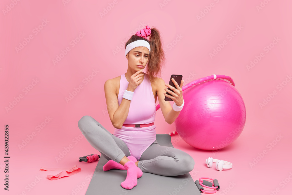 Displeased European woman looks at smartphone display has sporty figure checks messages or newsfeed in social networks sits on comfortable mat isolated over pink background. Sport activity concept Displeased European woman looks at smartphone display has sporty figure checks messages or newsfeed in social networks sits on comfortable mat isolated over pink background. Sport activity concept