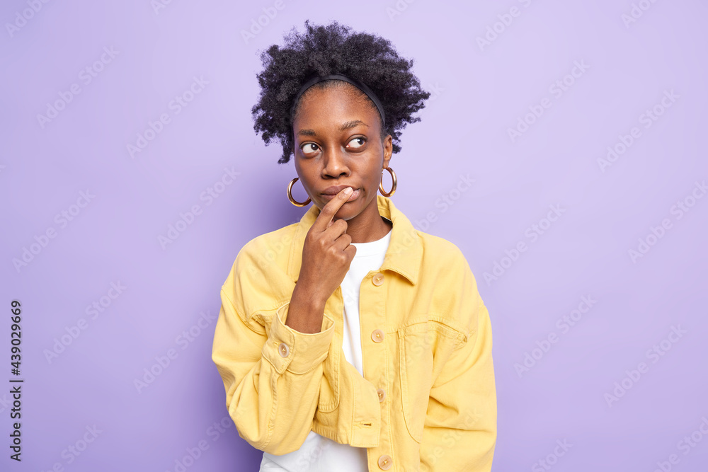 Horizontal shot of pensive dark skinned young woman makes choice keeps index finger on lips thinks deeply about something dressed in yellow jacket isolated over purple background. Deep thoughts Horizontal shot of pensive dark skinned young woman makes choice keeps index finger on lips thinks deeply about something dressed in yellow jacket isolated over purple background. Deep thoughts