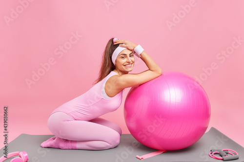 Sport at home during quarantine. Happy slim woman with pony tail leans on fitness ball takes break after workout leads active lifestyle dressed in body suit isolated over pink studio background.