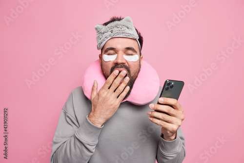 Photo of sleepy man covers mouth with hand wants to have rest scrolls social networks via smartphone applies patches to reduce puffiness under eyes wears sleepmask travel pillow around neck. © wayhome.studio 