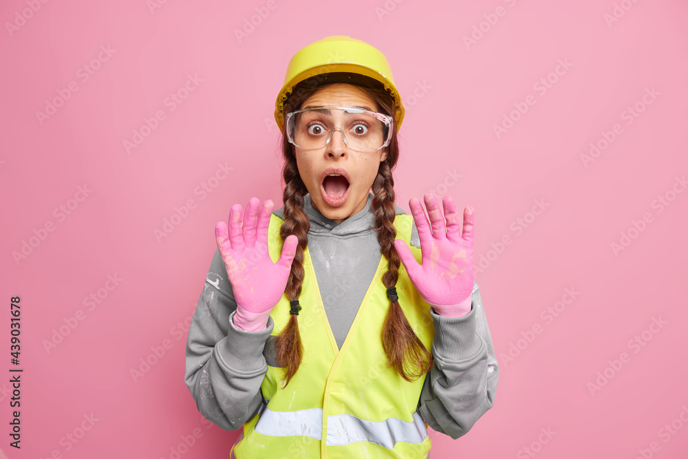 Scared woman industrial worker keeps palms raised with fearful expression wears protective helmet transparent glasses uniform isolated over pink background. Frightened female builder with pigtails Scared woman industrial worker keeps palms raised with fearful expression wears protective helmet transparent glasses uniform isolated over pink background. Frightened female builder with pigtails