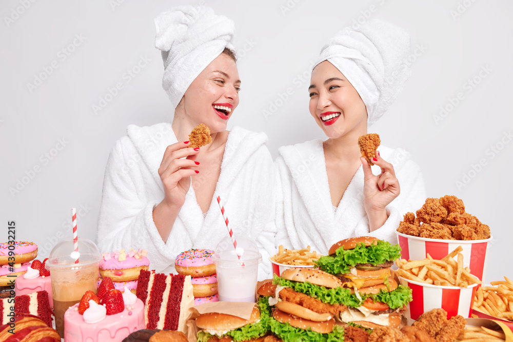 Two happy diverse women look gladfully at each other hold nuggets eat tasty fast food enjoy cheat meal dressed in casual domestic clothes isolated over white background. Unhealthy nutrition. Two happy diverse women look gladfully at each other hold nuggets eat tasty fast food enjoy cheat meal dressed in casual domestic clothes isolated over white background. Unhealthy nutrition.