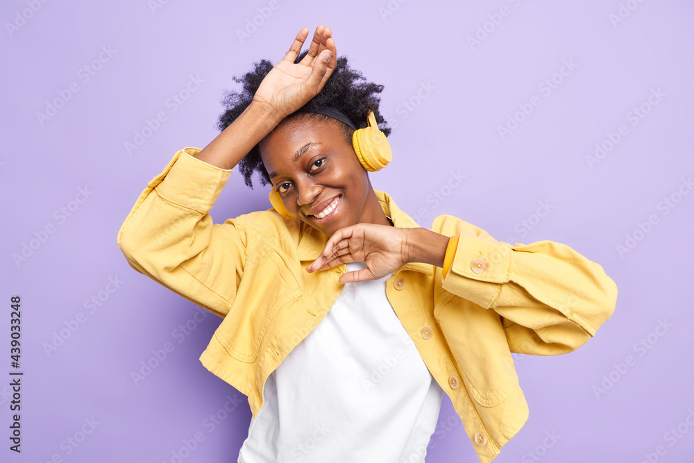 Glad dark skinned teenage girl raises arms smiles toothily has upbeat mood looks happily at camera listens music with wireless headphones wears fashionable clothes isolated over purple wall. Glad dark skinned teenage girl raises arms smiles toothily has upbeat mood looks happily at camera listens music with wireless headphones wears fashionable clothes isolated over purple wall.