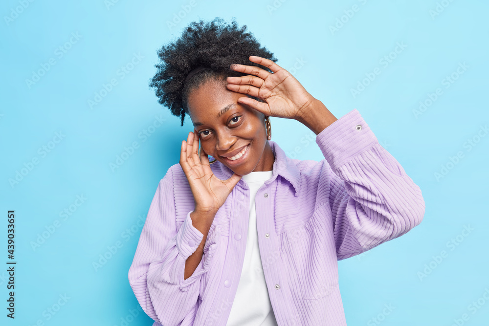Studio shot of glad dark skinned Afro American woman has gentle happy face expression feels shy looks directly at camera wears purple shirt isolated over blue background. Human emotions concept Studio shot of glad dark skinned Afro American woman has gentle happy face expression feels shy looks directly at camera wears purple shirt isolated over blue background. Human emotions concept