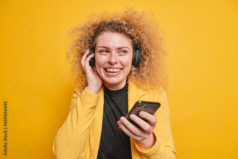 Image of cheerful curly woman uses modern technologies for listening music smiles toothily holds mobile phone wireless headphones on ears poses against yellow background. People leisure concept Image of cheerful curly woman uses modern technologies for listening music smiles toothily holds mobile phone wireless headphones on ears poses against yellow background. People leisure concept