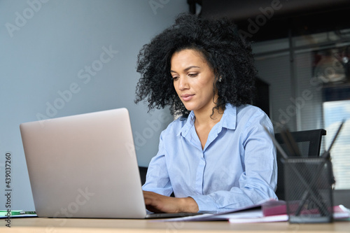 Young serious African American female ceo lawyer businesswoman sitting at desk working typing on laptop computer in contemporary corporation office. Business technologies concept.