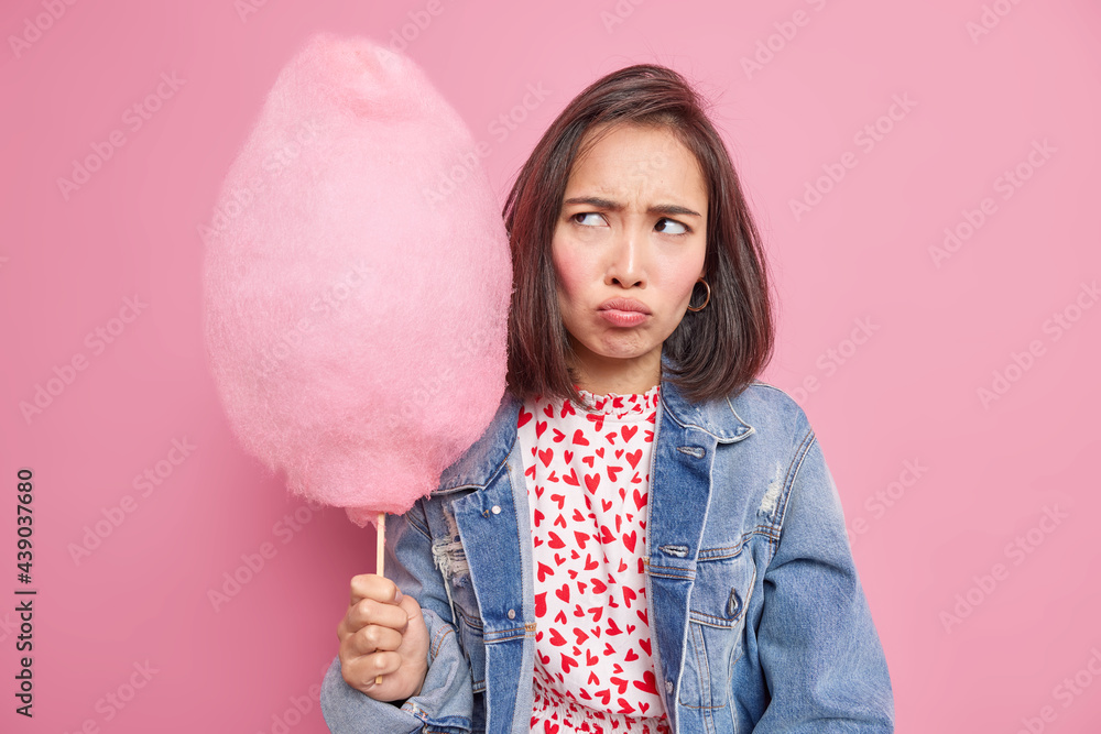 Dissatisifed sulking Asian teenage girl holds sweet cotton candy on stick doesnt like something wears denim jacket poses against pink background. Portrait of lovely female model with tasty dessert Dissatisifed sulking Asian teenage girl holds sweet cotton candy on stick doesnt like something wears denim jacket poses against pink background. Portrait of lovely female model with tasty dessert
