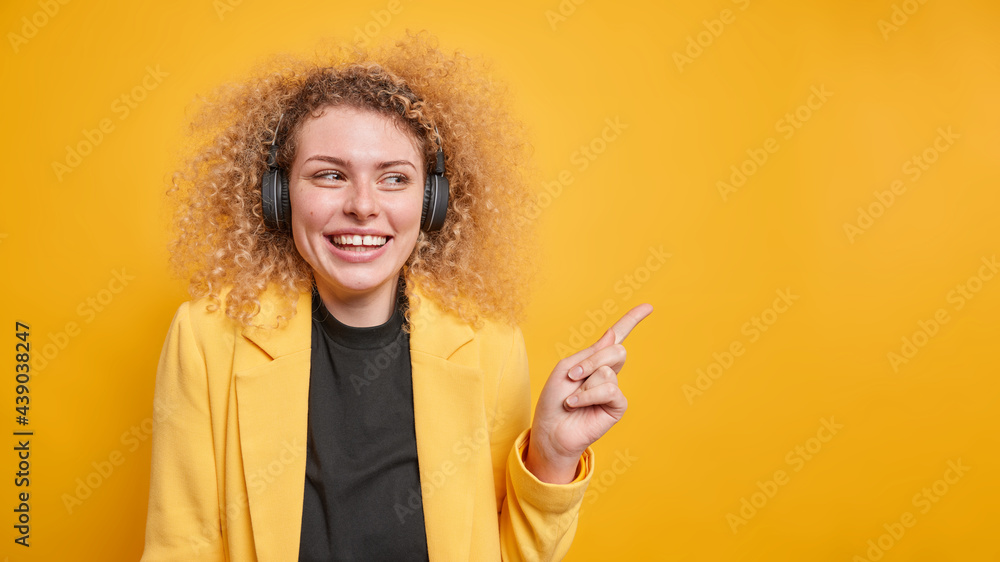 Studio shot of pleasant looking cheerful woman with curly hair wears black wireless headphones dressed in formal clothes indicates away on copy space isolated over yellow background. I advice this Studio shot of pleasant looking cheerful woman with curly hair wears black wireless headphones dressed in formal clothes indicates away on copy space isolated over yellow background. I advice this