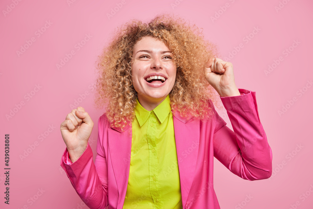 Happy upbeat curly haired young European woman has beaming toothy smile makes triumph dance clenches fists looks above dressed in green shirt pink formal jacket celebrates success poses indoor Happy upbeat curly haired young European woman has beaming toothy smile makes triumph dance clenches fists looks above dressed in green shirt pink formal jacket celebrates success poses indoor