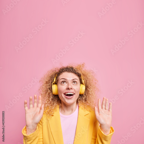 Glad woman with curly hair keeps palms forward camera focused above has cheerful expression wears formal yellow jacket poses against pink background blank space for your advertising content.