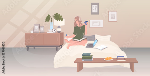 woman sitting on bed drinking coffee listening music writing report during coronavirus quarantine