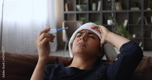 Indian frown unhealthy young woman lean on cushion of sofa having high fever put wet cloth on forehead holding electronic thermometer checks temperature feels unwell and weak, grippe symptoms concept