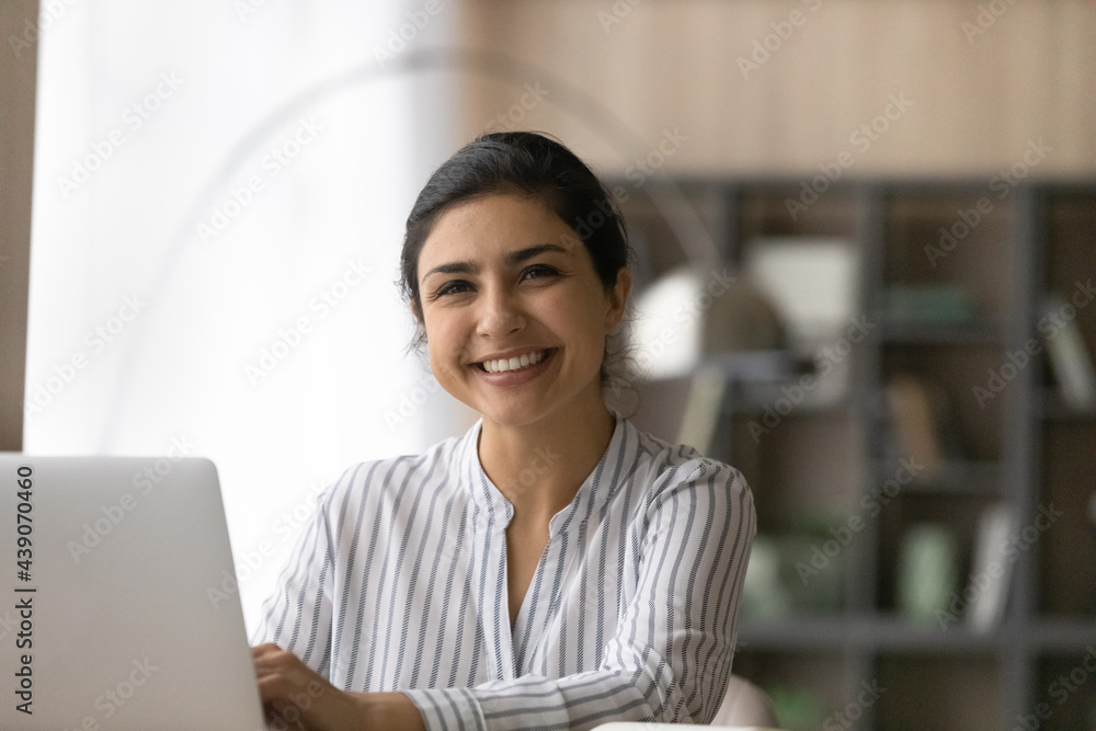 White collar. Happy young indian female accountant consultant sit at workplace by laptop take break in typing document look at camera. Headshot portrait of cheerful ethnic woman use pc in office work White collar. Happy young indian female accountant consultant sit at workplace by laptop take break in typing document look at camera. Headshot portrait of cheerful ethnic woman use pc in office work