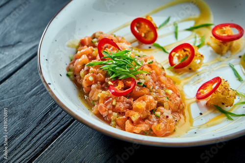 Delicious appetizer - salmon tartare with avocado and hot chilie pepper on a white plate on black wooden background.