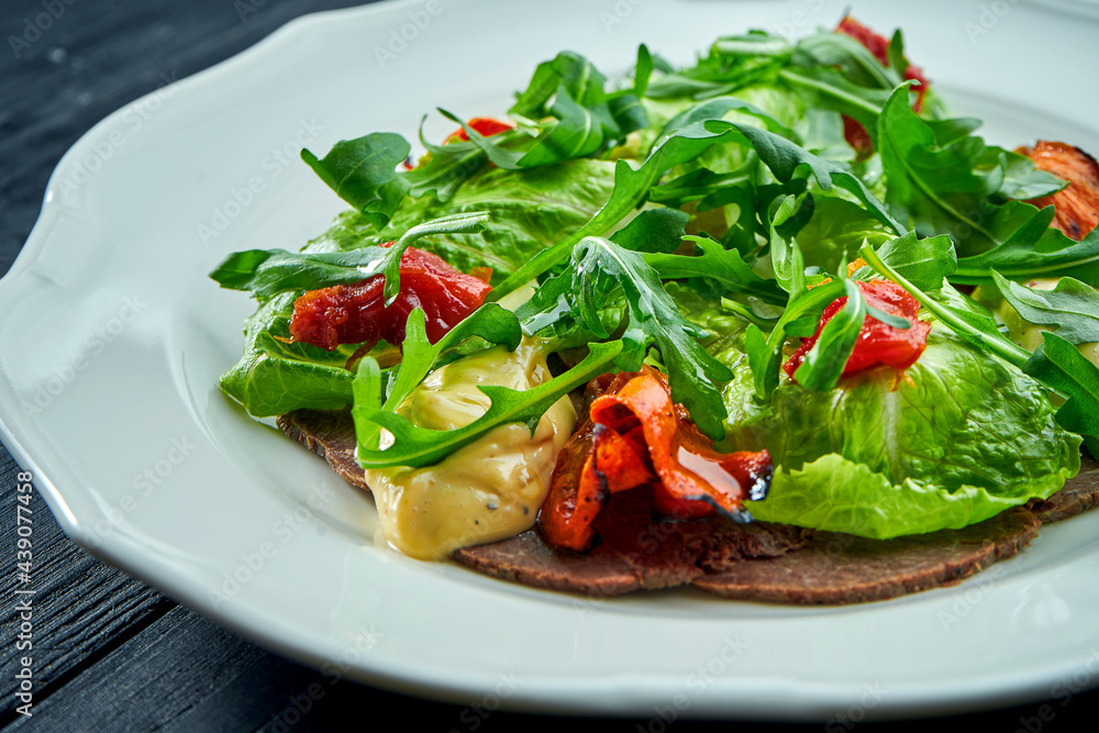 The traditional Italian appetizer salad is vitello tonnato. Thinly sliced beef with rucola, tomatoes and pesto, served in a white plate on a dark background