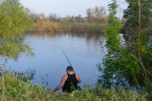 Fisherman sits with fishing rod on bank of the river. View from the back