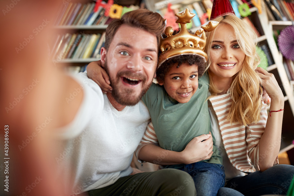 Fototapeta premium Happy couple taking a selfie with their adopted son at kids birthday party at home. Family, celebration, together