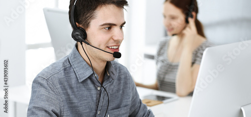 Casual dressed young man using headset and computer while talking with customers online. Group of operators at work. Call center