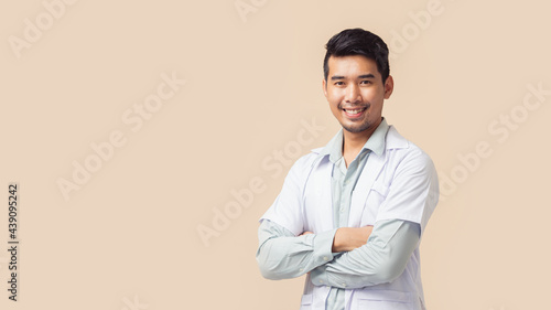 Asian man doctor in lab coat smiling with arm crossed with copy space on brown background.