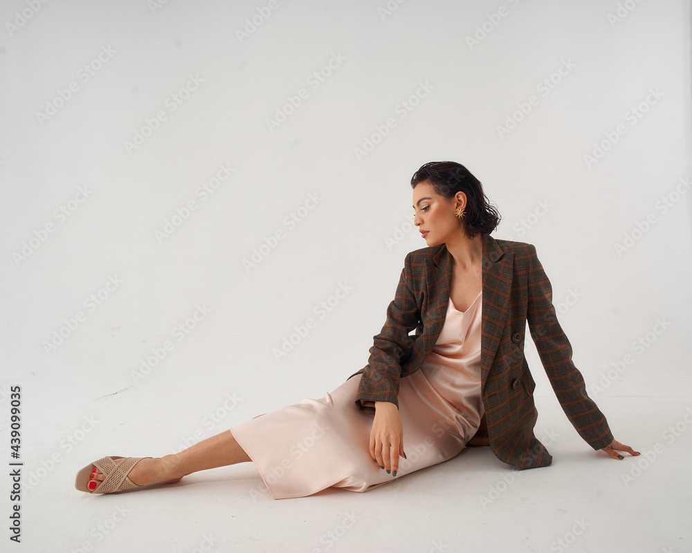 Elegant woman in peach color silk slip dress and brown squared wool blazer sitting and posing in studio at white background. Pretty stylish brunete girl with makeup and wet hair full length portrait Elegant woman in peach color silk slip dress and brown squared wool blazer sitting and posing in studio at white background. Pretty stylish brunete girl with makeup and wet hair full length portrait