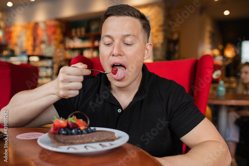 A young man puts berry from chocolate dessert in his mouth, tasting dessert at table in cozy cafe