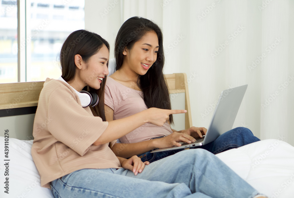 Young asian teen and her friend use laptop computer on the bed to relax in the bedroom, enjoy leisure weekend at home. Stress free concept