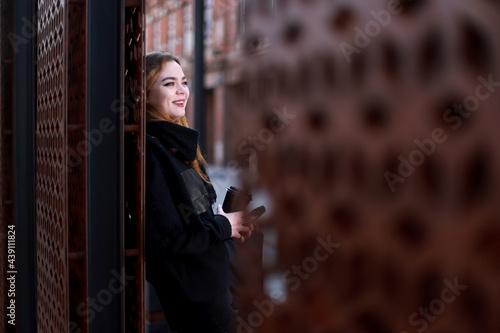 A beautiful model in black clothes with black coffee stands and laughs near a metal wall. Life style