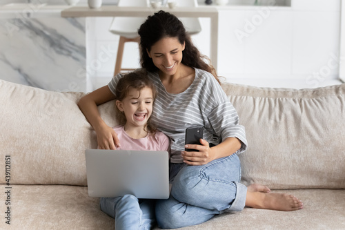 Excited loving Latino mom and little ethnic daughter have fun using modern electronic gadgets at home together. Smiling young Hispanic mother and small girl child browse web on cellphone laptop.