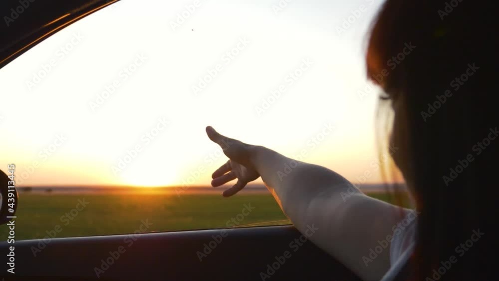 A young tourist girl waves her hand out the window. The teen travels at sunset and enjoys the ride by car.