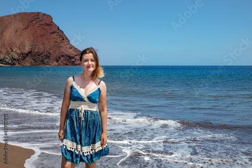 Happy millennial woman wearing a blue paisley dress while enjoying a beautiful day on La Tejita beach with views towards the frothy waves and volcanic Montana Roja in Tenerife, Canary Islands, Spain photo