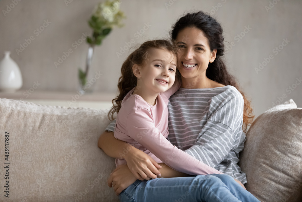 Portrait of smiling young Latino mother and little teen biracial daughter relax on sofa hug cuddle. Happy Hispanic mom and small ethnic girl child embrace show love care. Family unity concept. Portrait of smiling young Latino mother and little teen biracial daughter relax on sofa hug cuddle. Happy Hispanic mom and small ethnic girl child embrace show love care. Family unity concept.