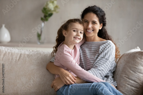 Portrait of smiling young Latino mother and little teen biracial daughter relax on sofa hug cuddle. Happy Hispanic mom and small ethnic girl child embrace show love care. Family unity concept.