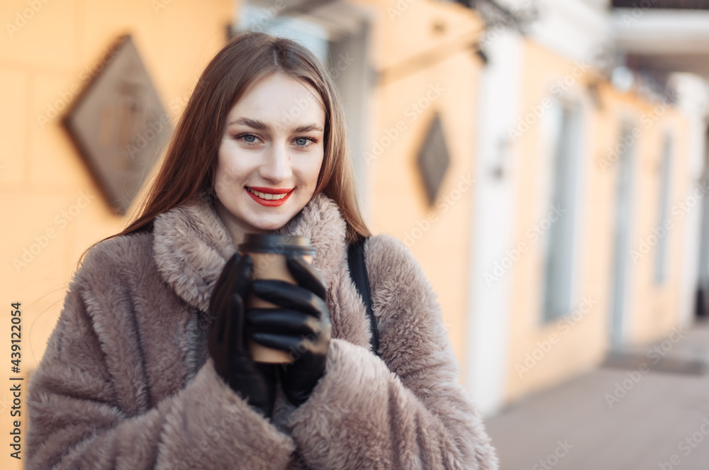 Young cheerful woman in a luxurious fur coat drinks coffee on the go in the city Young cheerful woman in a luxurious fur coat drinks coffee on the go in the city