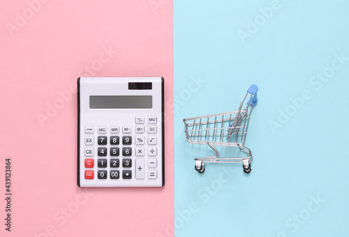 Calculator and shopping trolley on a blue-pink pastel background. Top view