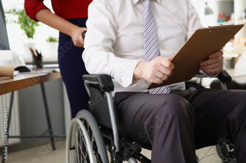 Woman secretary rolls CEO in wheelchair with documents and reports in hands