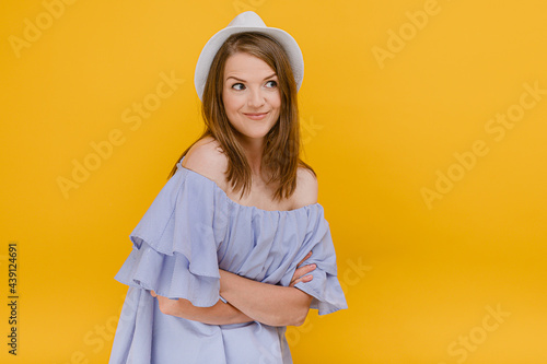 Beautiful woman in straw hat wear stylish summer dress with bare shoulders with arms folded looks outside away standing isolated against a yellow background