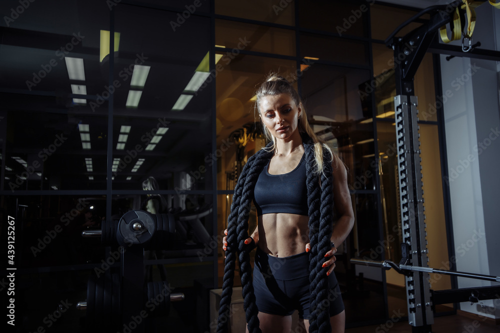 Sexy fit woman dressed in sportswear posing with battle ropes in dark fitness gym