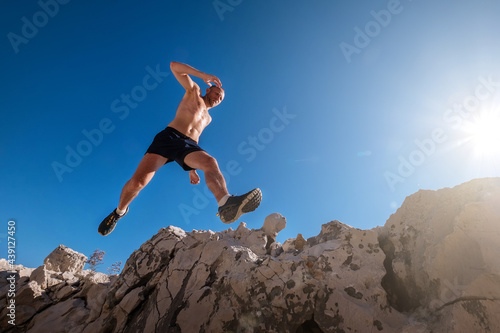 Active fast running mountain sweaty body muscular sky runner jumping over the cleft cliff during the morning jogging. Sporty people activities wide-angle concept image.
