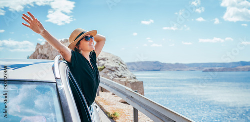 Cheerful Woman portrait enjoying the seaside road trip. Dressed a black dress, straw hat and sungllasses she wide opened arms and shining with happiness. Summer vacation traveling by auto concept.