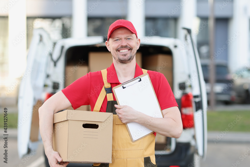 Male courier in uniform holds clipboard and cardboard box in background of truck with boxes Male courier in uniform holds clipboard and cardboard box in background of truck with boxes