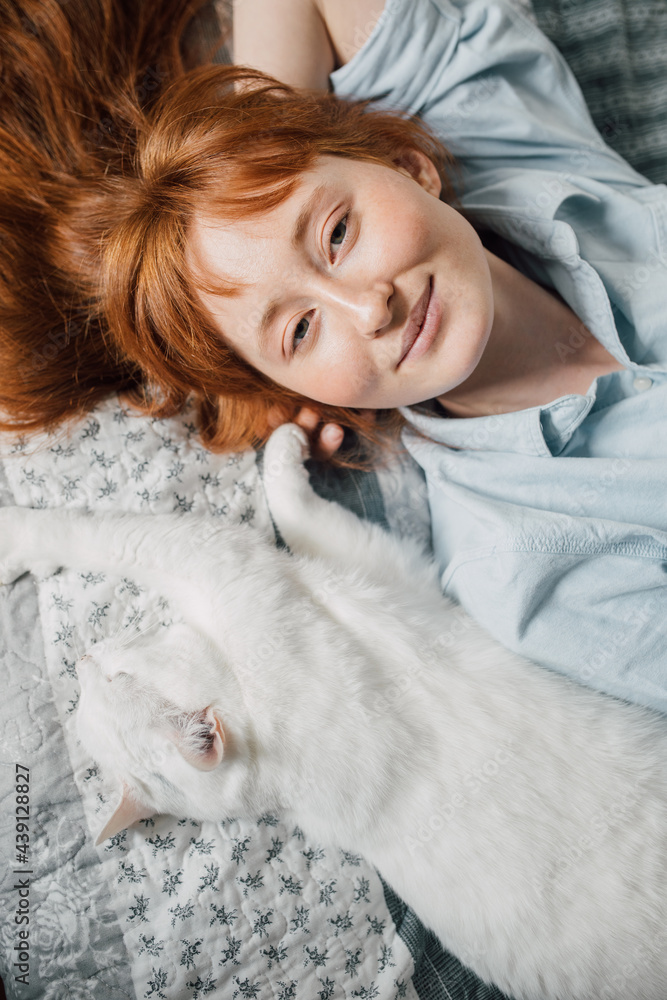 Woman looking at the camera and laying at the back at the bed near her white cat