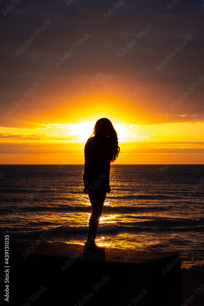 A gorgeous silhouette of a woman with long hair standing on the beach with the glow from the sun illuminating her outline as orange and yellow colors reflect off the water and cover the sky.