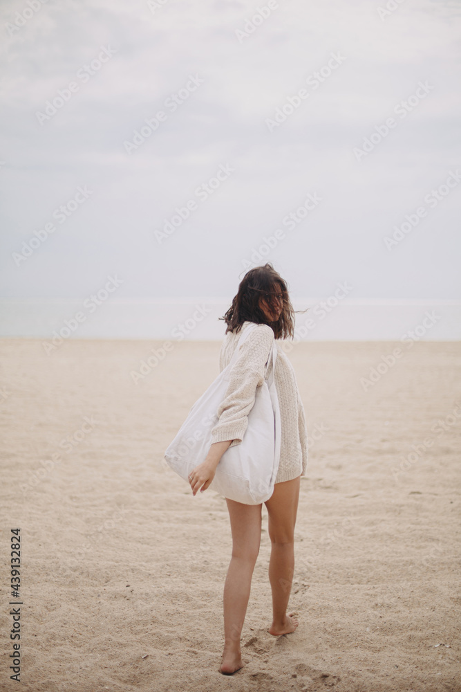 Beautiful stylish woman with windy hair and tote bag walking on sandy beach to sea, carefree moment. Stylish young female in knitted sweater enjoying vacation and relaxing on coast. Vertical image Beautiful stylish woman with windy hair and tote bag walking on sandy beach to sea, carefree moment. Stylish young female in knitted sweater enjoying vacation and relaxing on coast. Vertical image