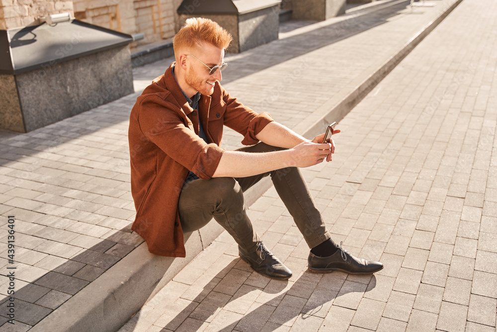 Man wearing sunglasses sitting in the rays of the sun and looking at his smartphone Man wearing sunglasses sitting in the rays of the sun and looking at his smartphone