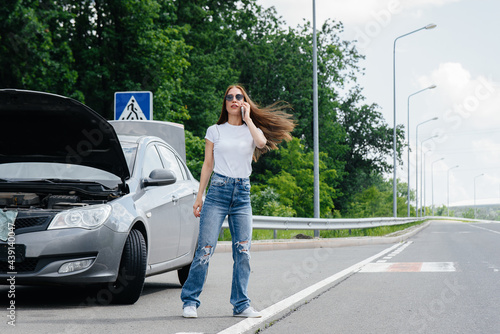 A young girl stands near a broken car in the middle of the highway and calls for help on the phone, while trying to stop passing cars. Failure and breakdown of the car. Waiting for help. © Andrii
