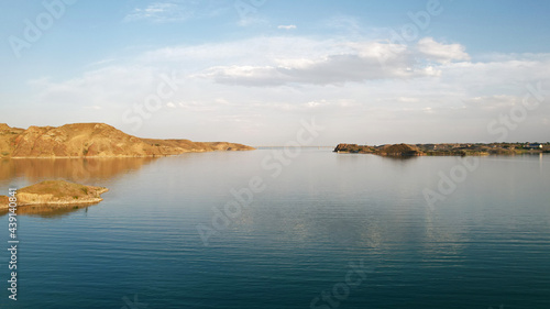 Flying over a lake with islands and rocks. Green water reflects the rays of the sun. Trees and bushes grow on the rocky ground, and houses are built. White clouds. Kapchagai Reservoir, Kazakhstan.